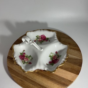 May include: A white, three-sectioned serving dish with a handle, featuring a floral rose design in pink and red. The dish has a gold trim and is placed on a wooden surface. Ideal for serving snacks or appetizers.