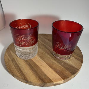 以下が含まれることがあります： 名前と日付が刻まれた、赤色のガラス製タンブラー2個。「Petoskey Sept 7 99」と「Ralph 1990」と書かれています。グラスは透明な模様のベースで、木製のサービングボードの上に置かれています。