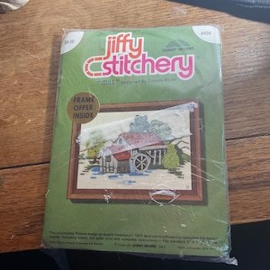 May include: A Jiffy Stitchery "Mill" cross-stitch kit. The kit features a framed image of a mill with a water wheel, trees, and a red-roofed building. The kit includes a printed design, wool yarns, and instructions.