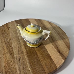 Op de afbeelding: Een kleine keramische theepot met een geel en wit kleurverloop, voorzien van een goudkleurige rand op het deksel en de tuit. Het lichaam van de theepot is versierd met een gedetailleerd reliëf van een donkere draak die eromheen kronkelt.