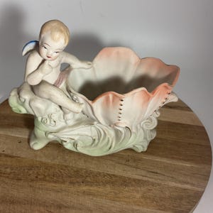 May include: A vintage ceramic planter featuring a cherub figure with a blue wing, seated on a floral base. The planter has a petal-shaped bowl with a pink and white color scheme, and gold accents.