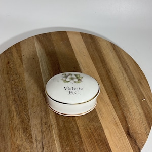 May include: A small, white ceramic trinket box with a gold rim. The lid features a floral design and the text "Victoria B.C." in black lettering. The box is round and sits on a wooden surface.