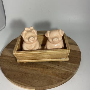May include: Two pink ceramic pig figurines sit inside a rectangular, light brown, wooden-style container. One pig has a bow on its head. The container is on a round wooden cutting board.