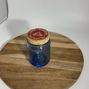 Könnte beinhalten: Ein blaues Vintage-Glas mit einem gold-roten Deckel. Der Deckel ist mit "Vicks VapoRub" und "Just Rub It On" beschriftet. Das Glas ist leer und steht auf einer Holzoberfläche. Das Glas hat ein klassisches Design.