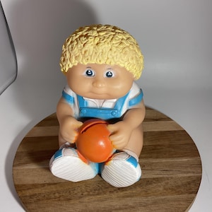 May include: A vintage Cabbage Patch Kid piggy bank with blonde hair, wearing a blue and white outfit, holding an orange basketball.