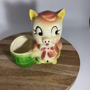 May include: A vintage ceramic planter in the shape of a pig. The pig has a cream-colored face with black eyes, a pink nose, and a red bow. It holds a green basket. The planter is on a wooden surface.
