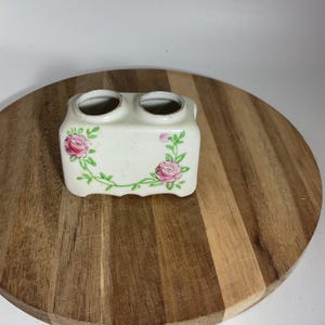 Puede incluir: Un pequeño recipiente de cerámica blanquecina con dos aberturas circulares. El recipiente está decorado con rosas rosas y vides verdes. La pieza es probablemente vintage y adecuada para un baño o tocador.
