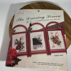 May include: A complete Christmas cross-stitch kit titled "The Dressing Screen." The kit features three red frames with cross-stitched images of musical instruments and notes. The kit includes frames, aida, needle, floss, and instructions. The package is marked "Made in America."