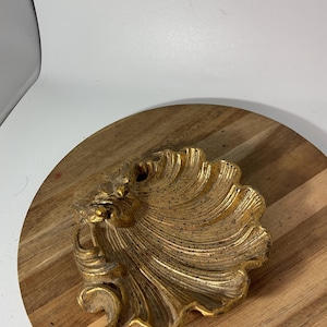 May include: A gold-colored decorative dish shaped like a seashell with a textured, ribbed surface and a distressed finish. The dish features a prominent, raised hinge area at the top and a scalloped edge, showcasing a metallic sheen throughout.