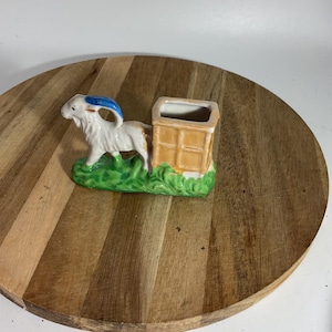 May include: A ceramic ornament depicting a white goat with blue horns and a tan body, standing on a green base. The goat is next to a tan rectangular structure. The ornament is on a round wooden surface.