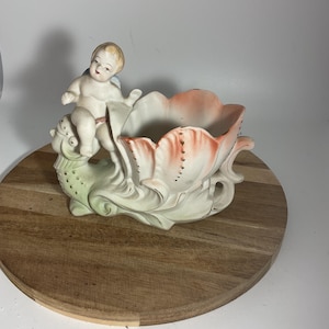 May include: A vintage ceramic planter featuring a cherub figure perched on a stylized floral design. The planter has a peach and white color scheme with gold accents, and is set on a wooden surface.