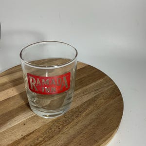 May include: A clear, cylindrical glass with the red text "Ramada Inn" printed on the front. The glass is on a round wooden surface. The glass is empty.