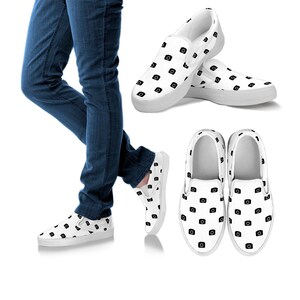 Pode incluir: Sapatos brancos de lona sem cadarços com um padrão de câmera preto. Os sapatos são usados por uma pessoa que usa calças jeans azuis.