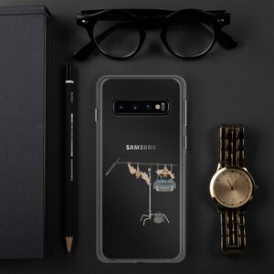 Può includere: Custodia trasparente per telefono Samsung con un'illustrazione di pipistrelli su un set cinematografico. Accanto, un orologio d'oro, occhiali neri, una matita e un taccuino nero su una superficie scura.