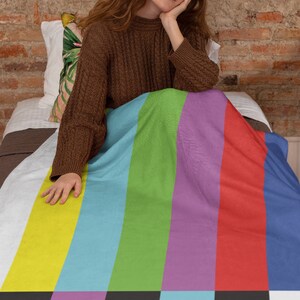 Puede incluir: Una mujer sentada en una cama con una manta de rayas de colores. La manta tiene un patr&oacute;n de rayas verticales en amarillo, azul, verde, morado, rojo y azul. La manta tiene un patr&oacute;n de prueba de televisi&oacute;n en blanco y negro en la parte inferior.