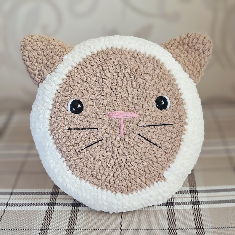 Peut inclure: Un coussin en forme de chat crochet&eacute; en marron et blanc avec des moustaches et des yeux brod&eacute;s en noir. Le coussin a un nez rose.