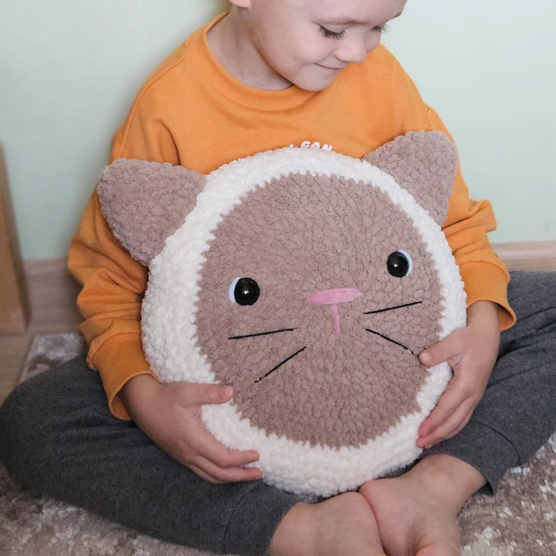 Peut inclure: Un enfant tient un coussin en forme de chat crochet&eacute; en marron et blanc. Le coussin a des yeux brod&eacute;s noirs, un nez rose et des moustaches.