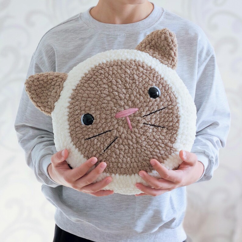 Peut inclure: Un coussin en forme de chat crochet&eacute; en marron et blanc avec un nez rose et des moustaches brod&eacute;es noires. Le coussin est tenu par une personne.