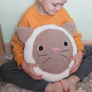 Peut inclure: Un enfant tient un coussin en forme de chat crochet&eacute; en marron et blanc. Le coussin a des yeux brod&eacute;s noirs, un nez rose et des moustaches.