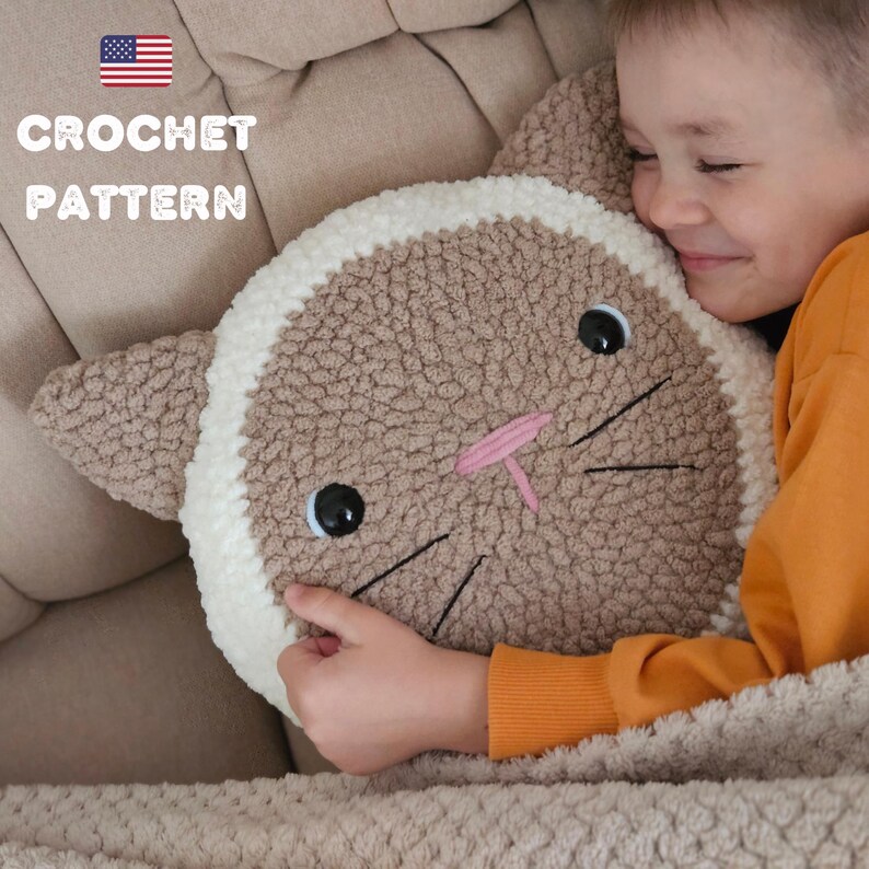 Peut inclure: Un coussin en forme de chat crochet&eacute; avec un motif brun et blanc. Le coussin a une bordure blanche et des yeux et des moustaches noirs brod&eacute;s. Un enfant tient le coussin et sourit.