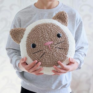 Peut inclure: Un coussin en forme de chat crochet&eacute; en marron et blanc avec un nez rose et des moustaches brod&eacute;es noires. Le coussin est tenu par une personne.
