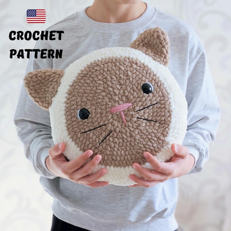 Peut inclure: Un mod&egrave;le de crochet pour un coussin de chat brun et blanc. Le coussin est rond et a une bordure blanche. Le chat a des yeux noirs, un nez rose et des moustaches noires.