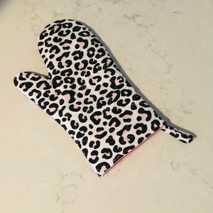 May include: A white oven mitt with a black and pink leopard print design. The mitt has a hanging loop.