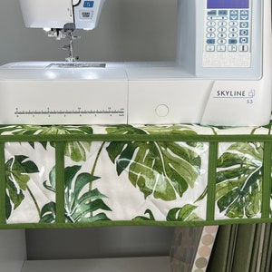 May include: A white sewing machine with a green and white fabric organizer with clear pockets. The sewing machine has a digital display and the text "SKYLINE S3" on the front.