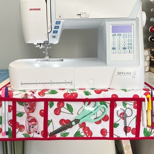 May include: A white sewing machine with a red and white cherry patterned fabric organizer with pockets for storing sewing supplies. The sewing machine has a digital display and the text "JANOME SKYLINE S3" on the front.