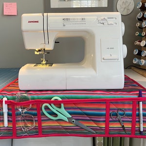 May include: A white Janome HD-3000 sewing machine sits on a colorful striped quilted sewing machine table with pockets for scissors, pens, and glasses.