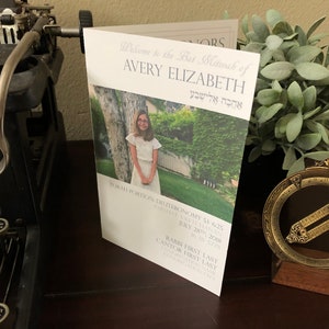 May include: A folded white program for a Bat Mitzvah featuring a photograph of a young person in a white dress. The text reads: Welcome to the Bat Mitzvah of AVERY ELIZABETH, TORAH PORTION: DEUTERONOMY 3:23-7:11, JULY 28TH, 2018, 16 AV 5778.