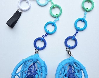 May include: Two blue and white macrame pacifier clips with a variety of blue rings and a blue woven oval-shaped pendant. Each clip has a black strap and a silver-colored metal clasp. The clips are designed to attach to a baby's clothing.
