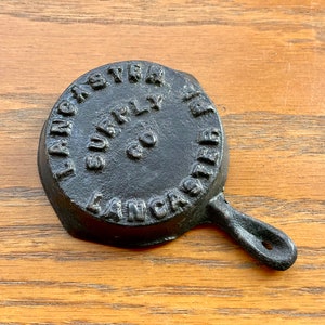 May include: A small black cast iron skillet with a handle. The skillet is embossed with the text "Lancaster Surly Co. Lancaster".