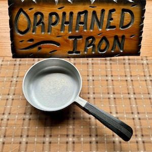 May include: A silver aluminum saucepan with a black plastic handle. The saucepan is on a brown and white checkered surface.