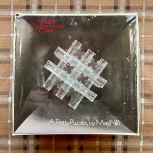 May include: A clear plastic puzzle in a white box with the text "Third Dimension One" and "A Pretty Puzzler, by MagNiF!" on the front.