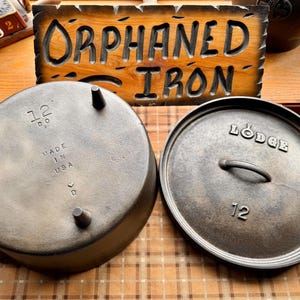 May include: A cast iron Lodge Dutch oven with a lid, both with the number 12. The pot has three legs and the lid has a handle. The words "Made in USA" are stamped on the pot. A wooden sign behind the pot reads "Orphaned Iron."
