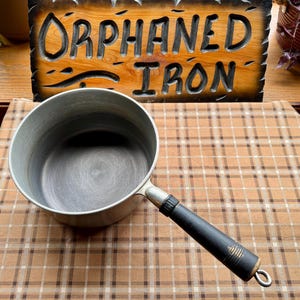 May include: A vintage metal saucepan with a black handle and a silver-colored body. The handle has a loop at the end. The sign in the background reads "Orphaned Iron" in carved wooden letters.