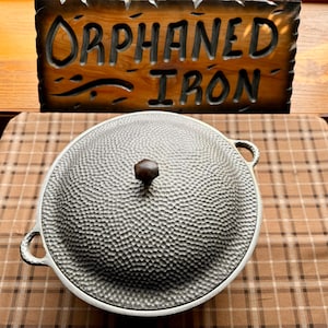 May include: A large, silver, cast iron pot with a domed lid and two handles. The pot has a hammered texture and a wooden knob on the lid. The pot is sitting on a plaid tablecloth.