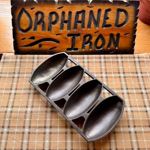 May include: A black cast iron corn stick pan featuring four elongated, oval-shaped molds arranged in a rectangular frame. The pan has a smooth, dark metallic finish and a sturdy construction with open spaces between each individual mold section.