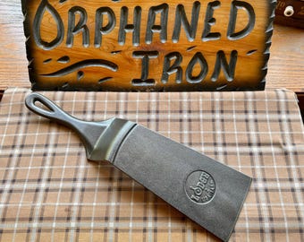 May include: A cast iron Lodge spatula with a rectangular blade and a handle featuring a circular cutout. The blade is embossed with the text Lodge 2 14SK USA. Behind the spatula is a wooden sign with the carved text ORPHANED IRON.