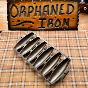 May include: A rectangular cast iron corn stick pan featuring five elongated, tapered molds for baking. The dark grey metal pan has a textured surface and small handles on each end. Behind the pan is a wooden sign with the text ORPHANED IRON.