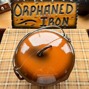 May include: A round, orange and brown enameled cast iron Dutch oven with a metal handle. The lid has a dark brown handle. The sign in the background reads "Orphaned Iron".
