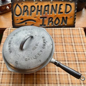 May include: A vintage cast iron skillet with a lid. The lid is marked "Wagner Drip Drop Skillet Cover No 9". The skillet has a black handle and a metal loop at the end. A wooden sign that says "Orphaned Iron" is in the background.