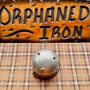 May include: A wooden sign with the words "ORPHANED IRON" carved into it, displayed above a metal object. The sign is brown with black lettering. The metal object is round and silver, with the word "WAGNER" on it.