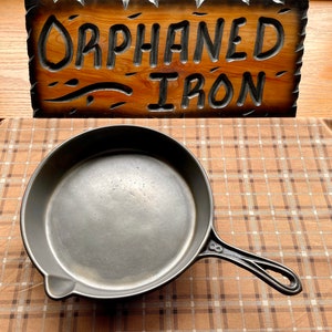 May include: A black cast iron skillet with a handle. The skillet is sitting on a brown and white checkered tablecloth. The skillet is empty.