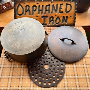 May include: An antique cast iron cooking pot with lid and trivet. The pot is marked "FAVORITE PIQUA WARE" and has the number "9" on the side. The lid has a handle. A wooden sign that says "ORPHANED IRON" is in the background.