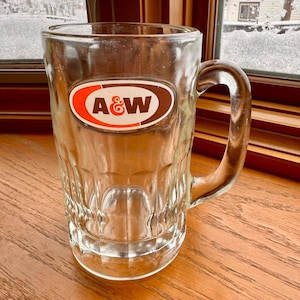 May include: A clear glass mug with a brown and white A&W logo on the side. The mug has a handle and is sitting on a wooden surface.
