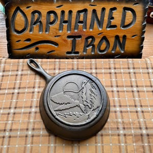May include: A black cast iron skillet featuring a raised relief design of a duck in flight over water with a sun in the background. The skillet has a single handle and is positioned in front of a wooden sign with the text ORPHANED IRON.