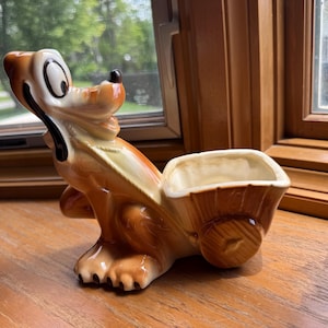May include: A ceramic figurine of a brown and tan dog with large eyes and a black nose, depicted in a seated position. The dog is attached to a hollow, cart-like container with a wood-grain texture, featuring a single wheel on the side.
