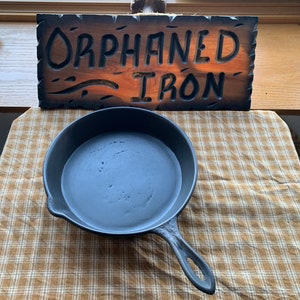 May include: A black cast iron skillet with a long handle. The skillet is sitting on a brown and white checkered tablecloth. A wooden sign with the words "Orphaned Iron" is in the background.